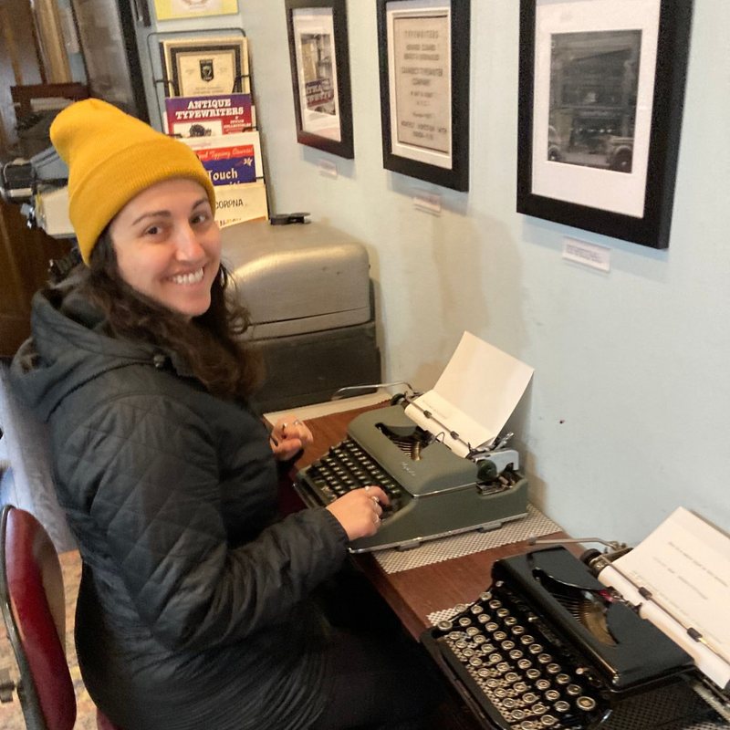 Sarah Fleming Steinberg in a yellow beanie, smiling over her shoulder while typing on a vintage typewriter